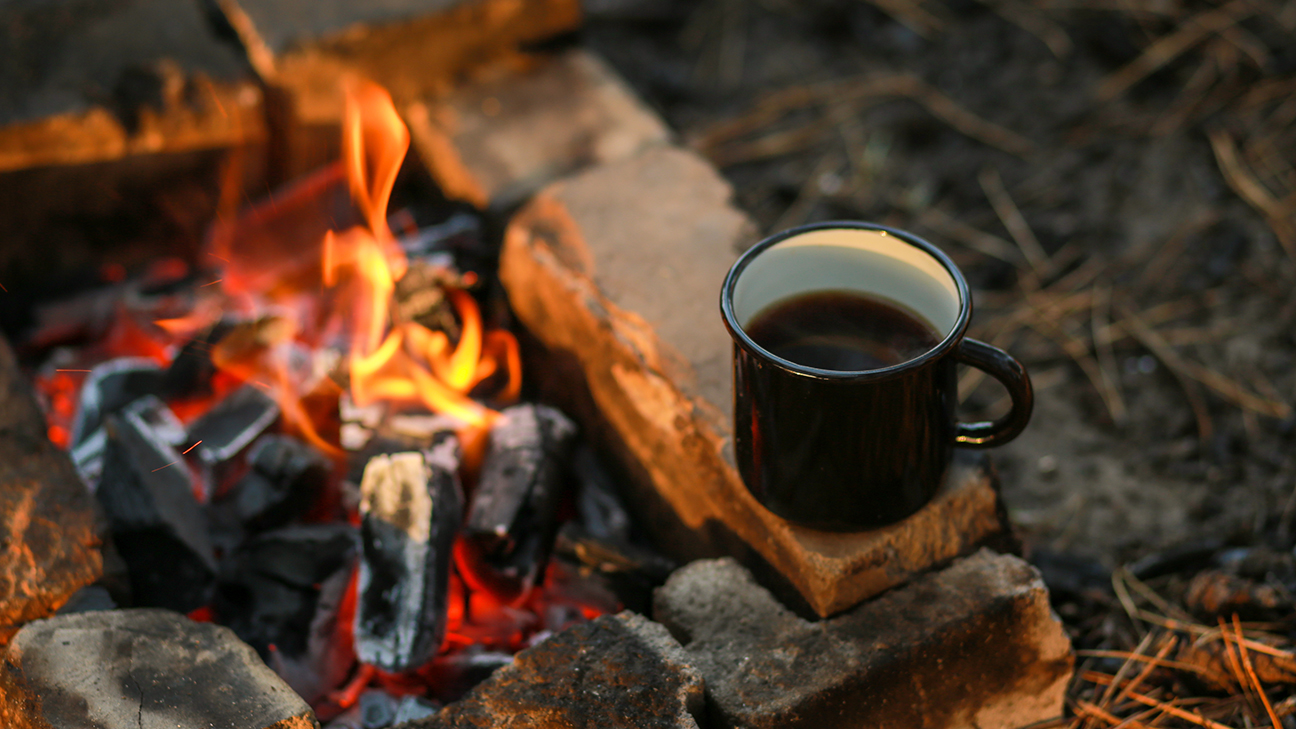 Black enamel with a cup of hot drink sits on a stone by the fire in the open air.